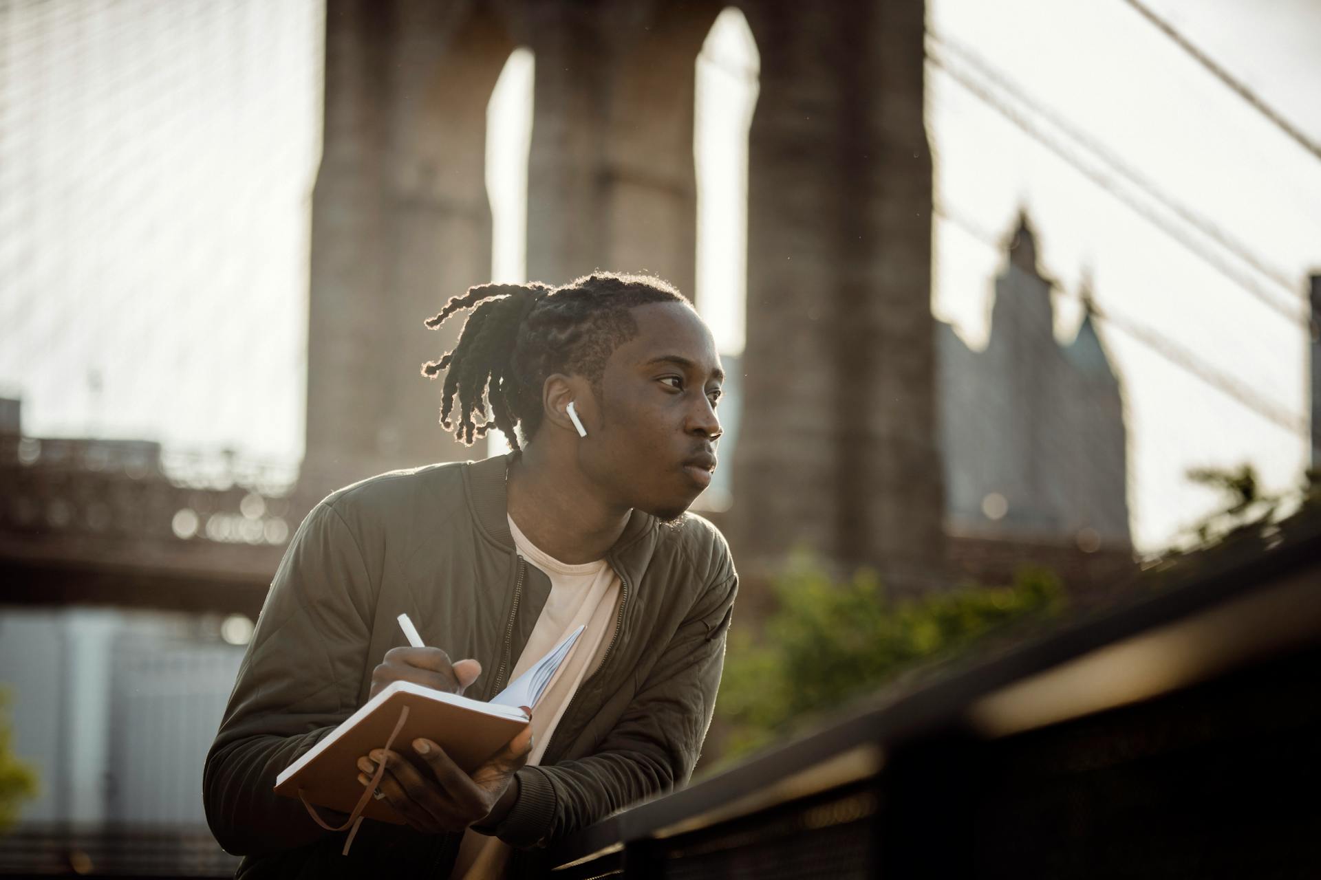 Positive black man listening to music with wireless earphones and taking notes in diary on street