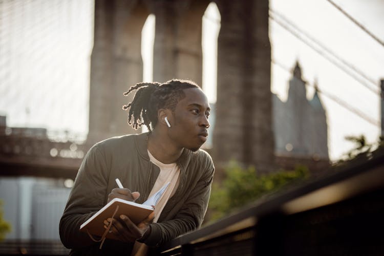 Positive Black Man Listening To Music With Wireless Earphones And Taking Notes In Diary On Street