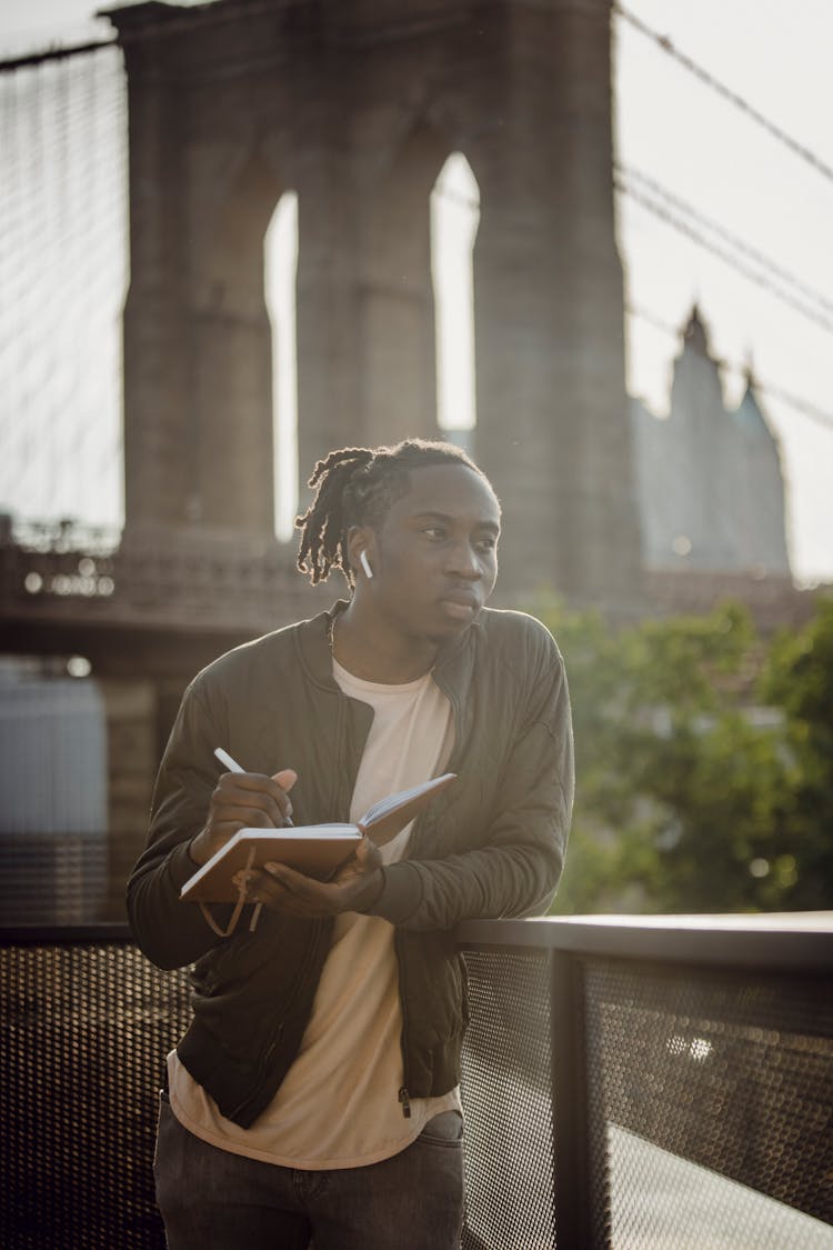 Young Black Man Painting Cityscape In Sketchbook While Standing On Embankment