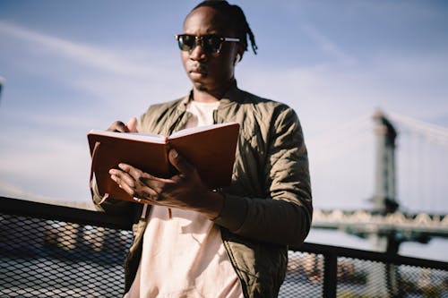 Free Black man in earphones writing in diary standing on bridge Stock Photo