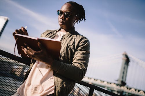 From below of adult African American guy in true wireless earphones and sunglasses standing with open notebook on city bridge under cloudy sky