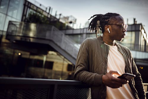 Free From below of dreamy African American male with pigtails and true wireless earphones standing on bridge with cellphone and listening to music while looking away Stock Photo
