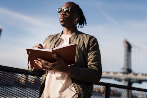 Stylish black guy in earphones with notebook standing on bridge