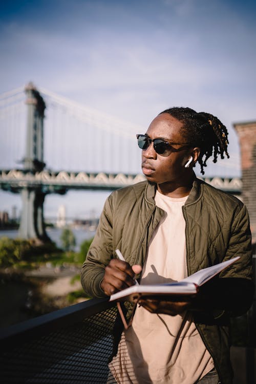 Young African American man in sunglasses listening information through wireless earphones and writing notes in notebook at sunny day