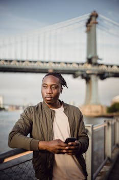 Young man using smartphone by bridge in urban setting, reflecting modern lifestyle.