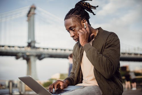 Free Low angle of concentrated black man in casual wear using laptop and earphones while working outdoors in park Stock Photo