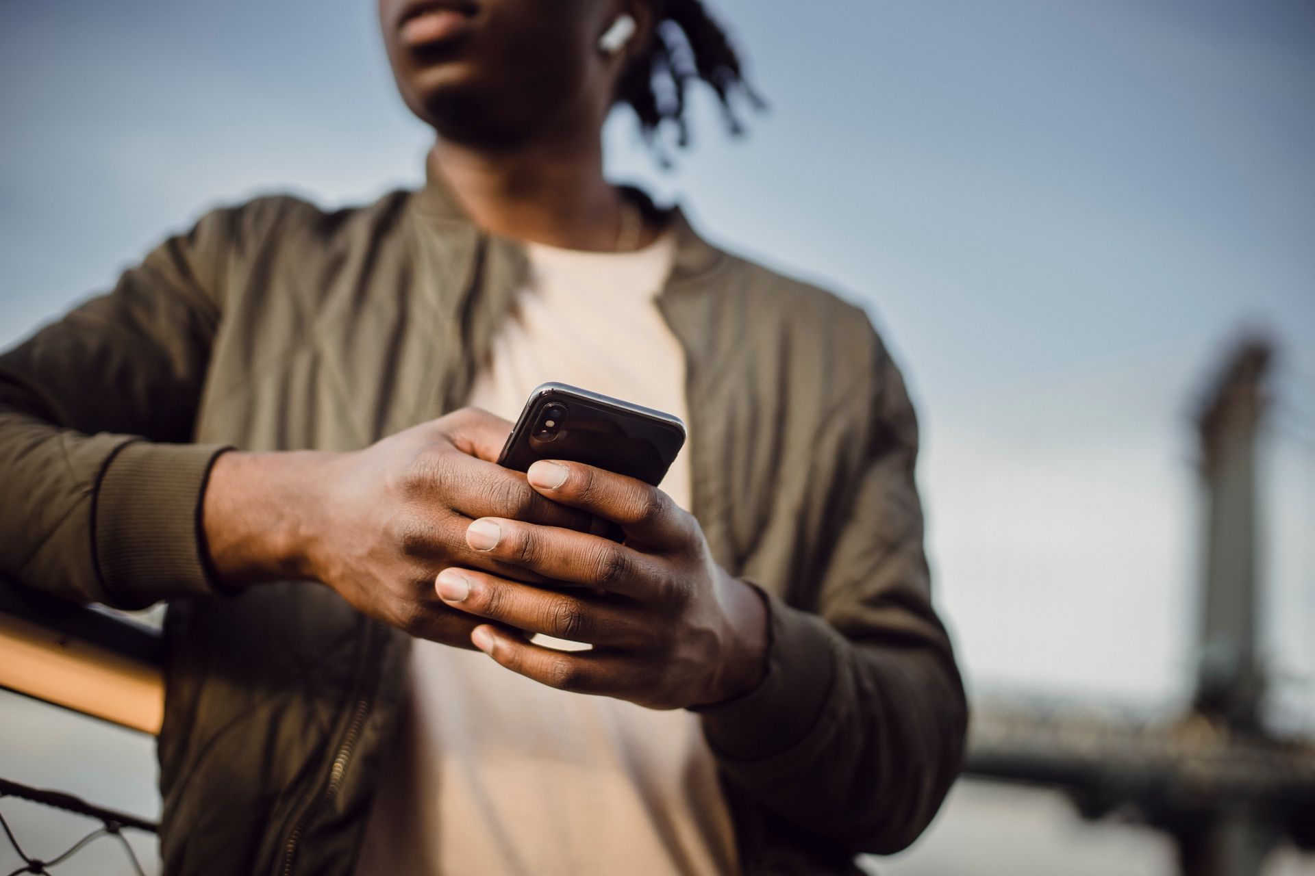 A man uses his smartphone outdoors in an urban environment, leaning on a railing with a cityscape backdrop.