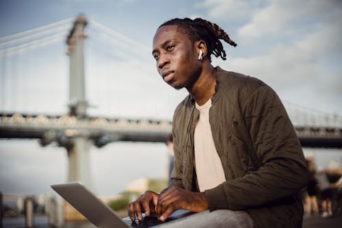 Low angle of concentrated black male freelancer working in city park while typing on keyboard and looking away