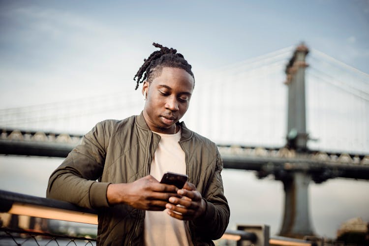 Focused Black Man Listening To Music In Earphones And Scrolling Smartphone