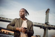 一个留着辫子的年轻人拿着智能手机，在桥边沉思着户外