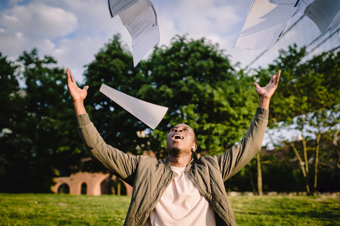 Free Cheerful young African American male student in casual clothes throwing college papers up in air while having fun in green park after end of exams Stock Photo