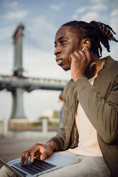 Low angle of serious young African American male listening to music through wireless earbuds while working on laptop against blurred backdrop of city bridge