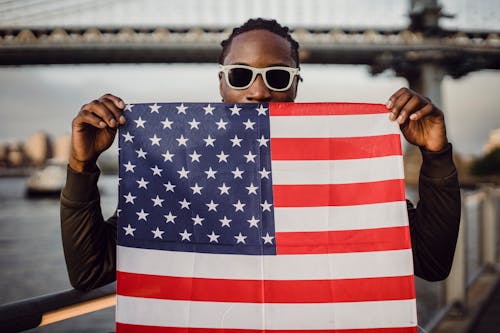 Young friendly black man in sunglasses with bandana with US flag print in hands standing against blurred Brooklyn Bridge in New York City