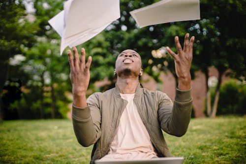 Free Content black remote worker throwing work papers in air happy to get rid of boring paperwork while sitting in green park with laptop Stock Photo