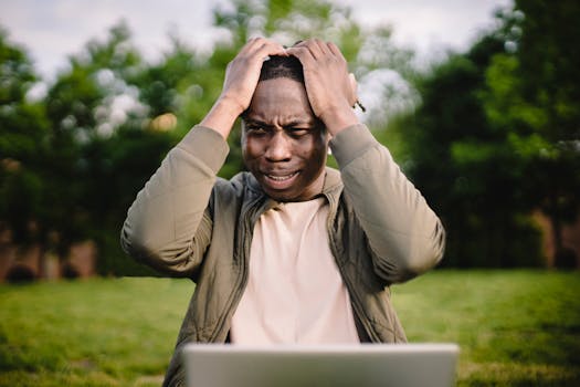 Man outdoors holding his head in frustration while using a laptop, reflecting stress or technical issues.