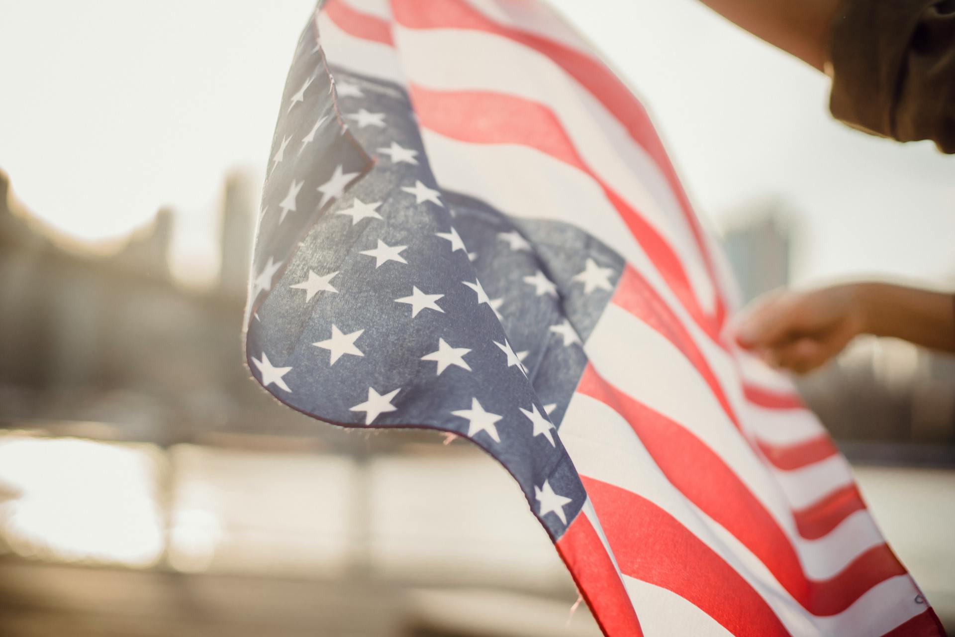 From below of crop person holding national flag of United States of America waving in wind on street against city river