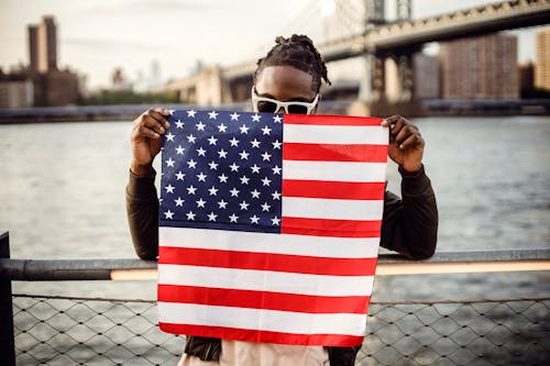 Ethnic male in casual clothes and sunglasses standing on embankment of city river while leaning on fence showing national flag of United States of America