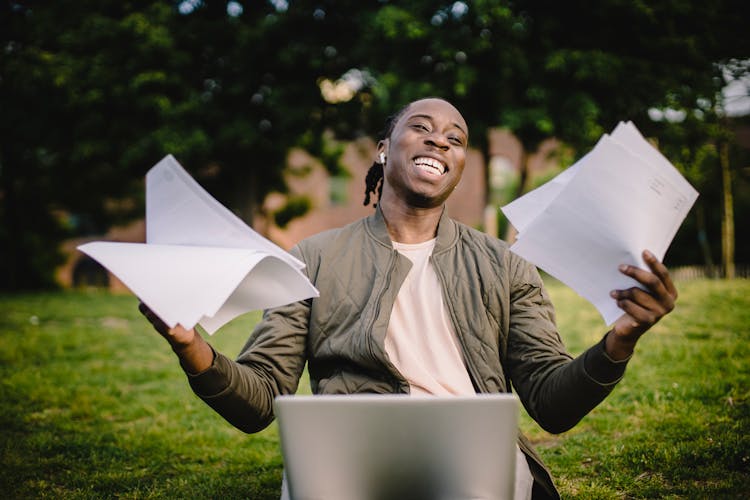 Happy African American Man Holding Sheets Of Paper In Front Of Laptop