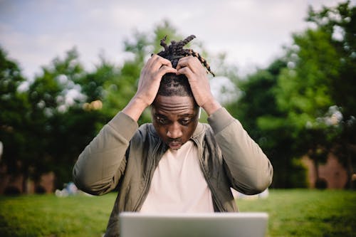 Free Upset young African American male freelancer clutching head with hands after failure in project while working remotely in park Stock Photo