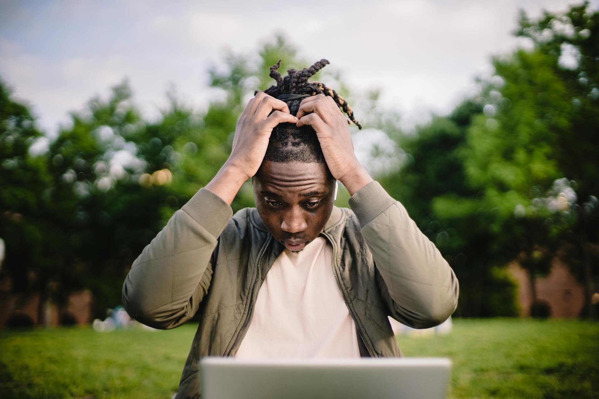 Upset young African American male freelancer clutching head with hands after failure in project while working remotely in park