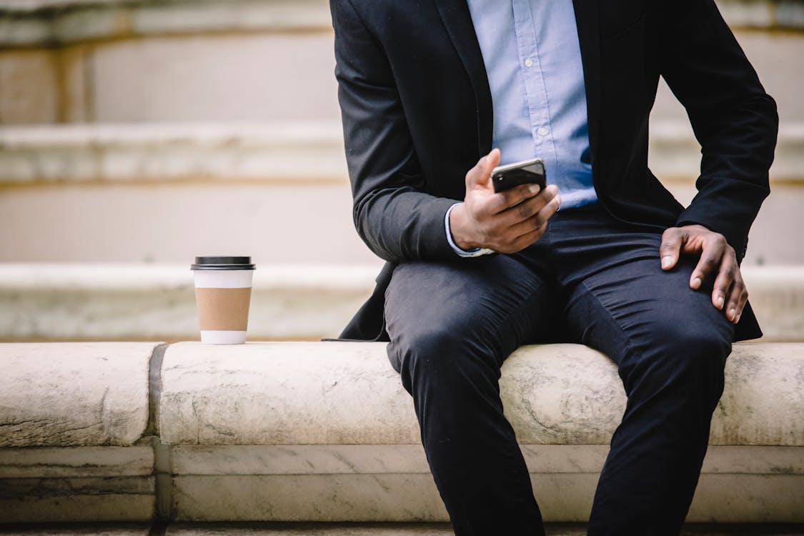 Crop faceless male manager in formal wear sitting on marble bench and messaging on mobile phone during coffee break