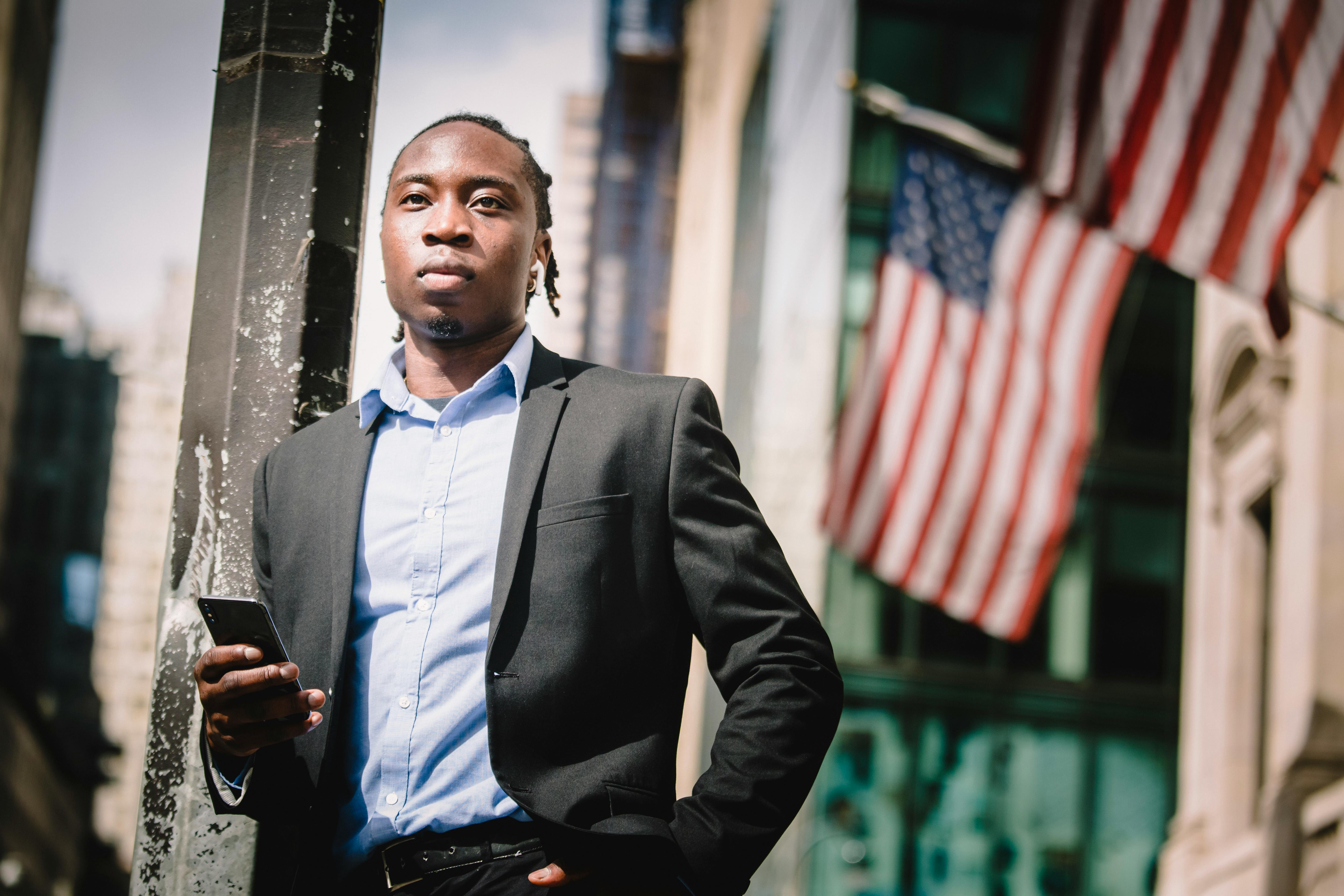 confident black businessman with smartphone against facade of building with us flags