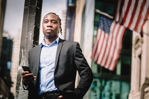 Low angle of serious well dressed ethnic male manager leaning on metal pillar on street and using cellphone against government building with hanging American flags