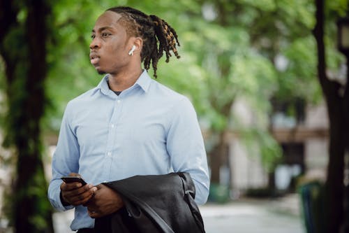 Stylish black businessman in TWS earbuds with smartphone waiting for meeting in park