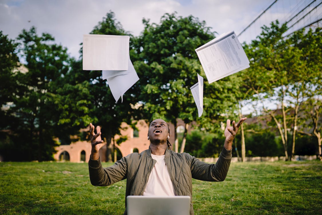 Happy young black male freelancer throwing papers while celebrating successful project during remote work in green park