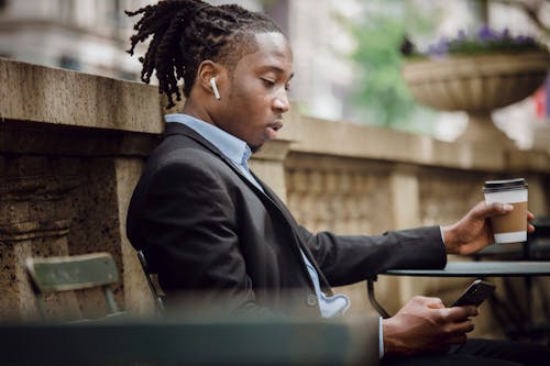 Free Serious black male messaging on smartphone while drinking coffee in outdoor cafe Stock Photo