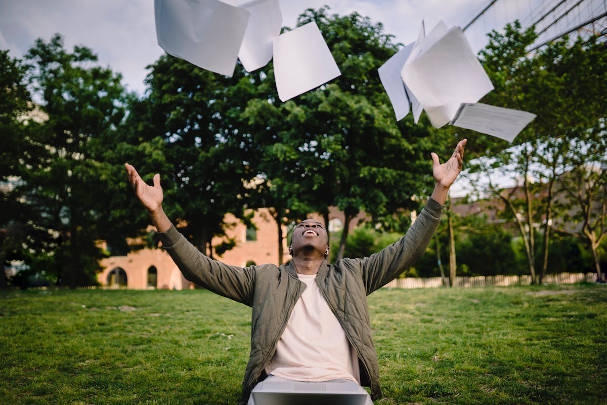 Happy young African American male student in casual outfit tossing university papers in air while having fun in green park after successfully completing academic assignments