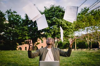 Overjoyed African American graduate tossing copies of resumes in air after learning news about successfully getting job while sitting in green park with laptop