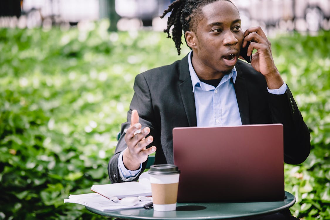 Free Young African American male entrepreneur in smart suit sitting in outdoor cafe with laptop and arguing on cellphone on sunny day Stock Photo