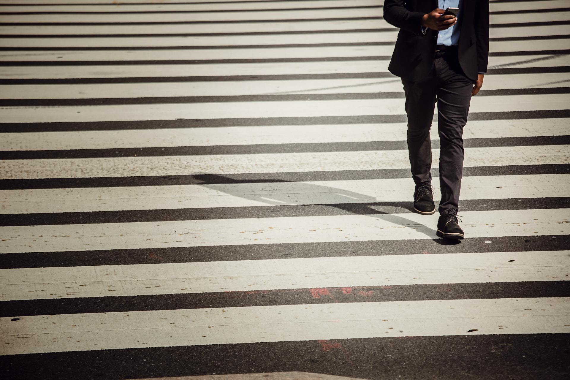 A businessman using a smartphone walks across a city crosswalk on a sunny day.