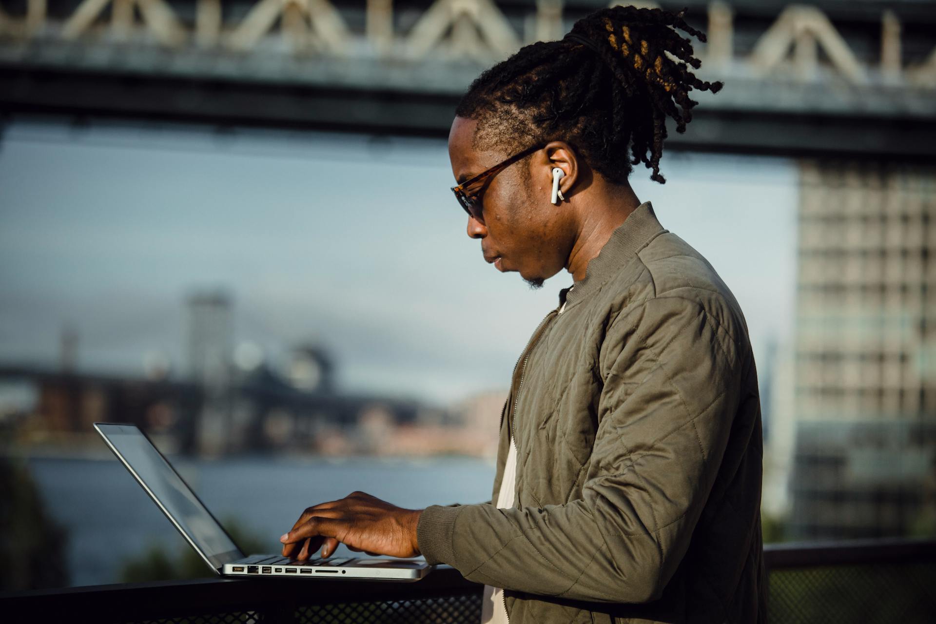 Side view of young African American male freelance worker in casual wear working on laptop and listening to music through true wireless earbuds against blurred river and city