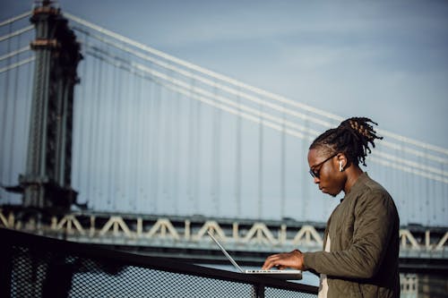 Free Side view of cool young African American man with dreadlocks in sunglasses and earbuds working on laptop on terrace railing against Manhattan Bridge and blurred blue sky Stock Photo