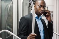 一位年轻英俊的非裔美国男性穿着正式西装，在通勤火车上用智能手机打电话