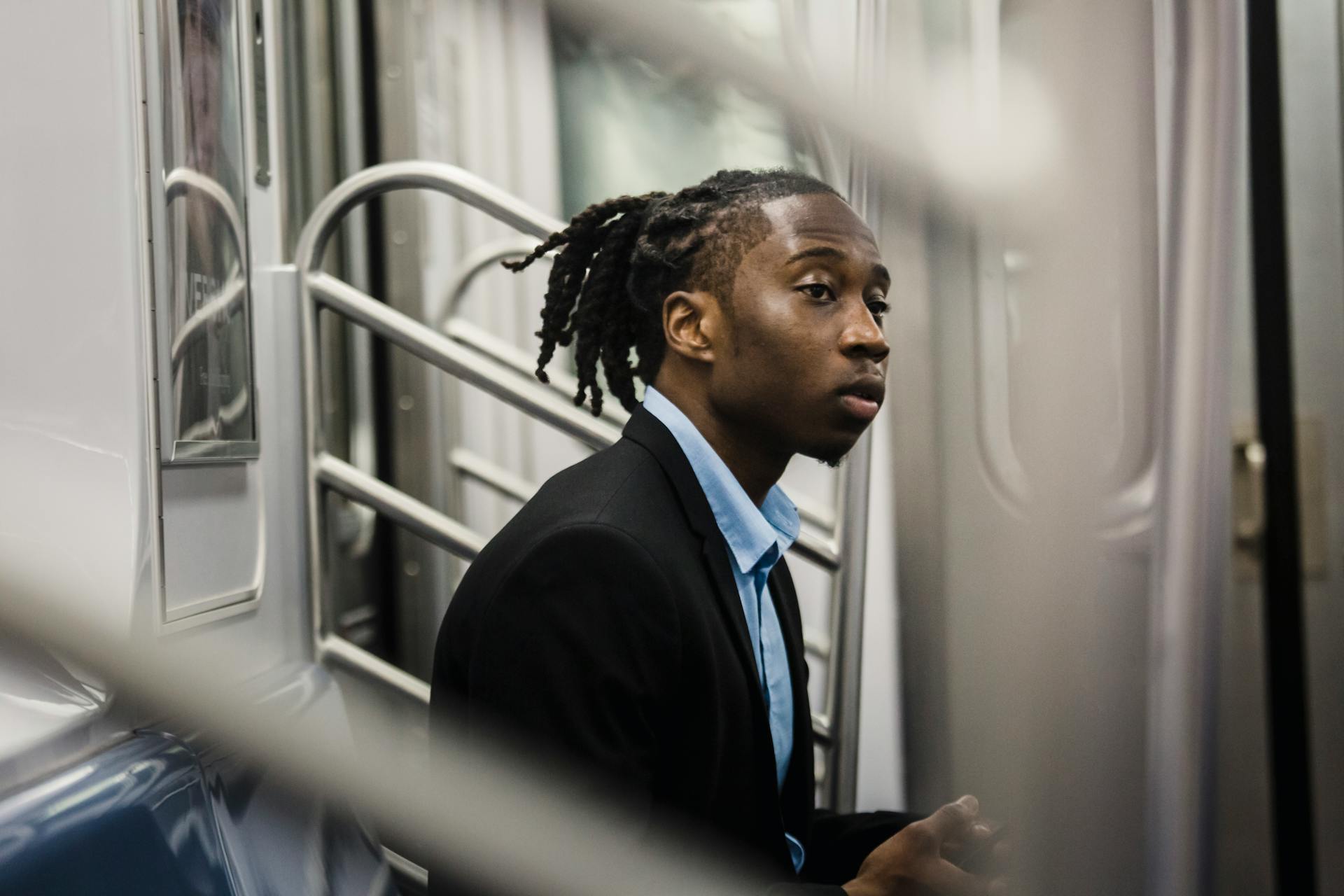 Side view of young stylish black man in formal wear with dreadlocks sitting on train seat and looking away with expression of slight interest on face