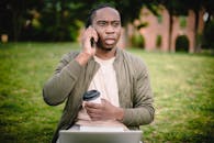 Young African American male in casual clothes with cup of coffee to go in hand talking on mobile with expression of displeasure and misunderstanding on face while sitting in park