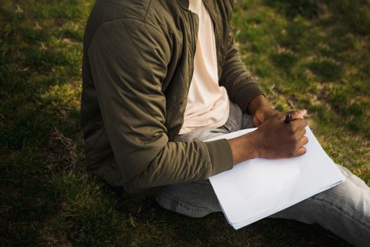 From above of crop African American male artist in casual clothes sitting on grass in park with clean sketch album and pen
