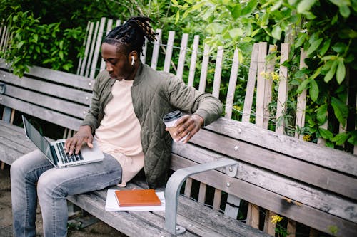 Free Young focused African American male student with coffee to go and notebooks working on laptop and listening to music through wireless earphones while sitting on wooden bench in green park Stock Photo