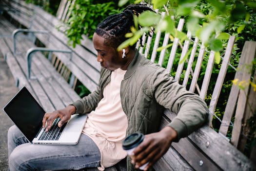 A young man enjoying remote work on his laptop with earbuds and coffee in a serene park setting.