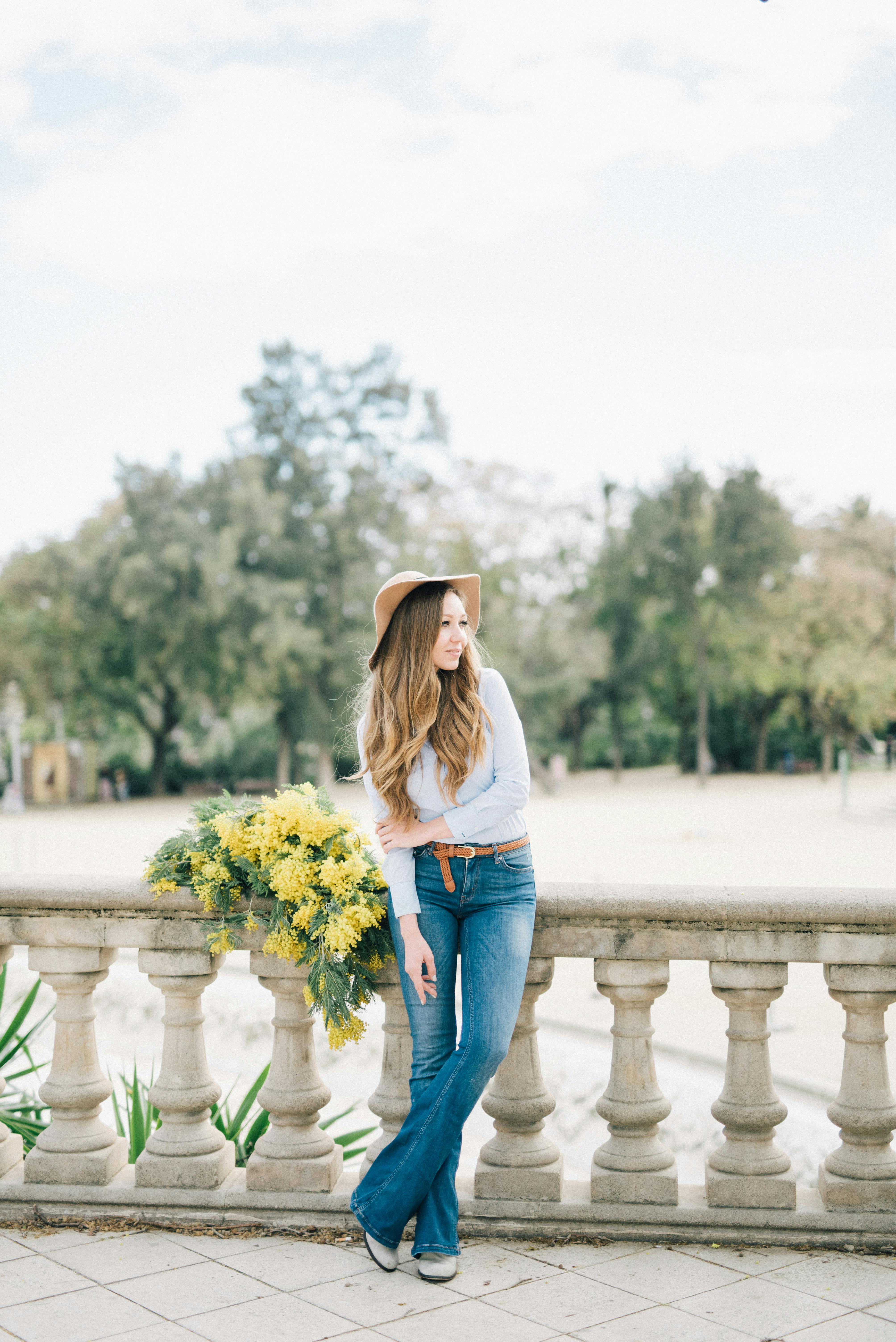 Stylish woman in denim and hat leaning on balusters with yellow flowers outdoors.