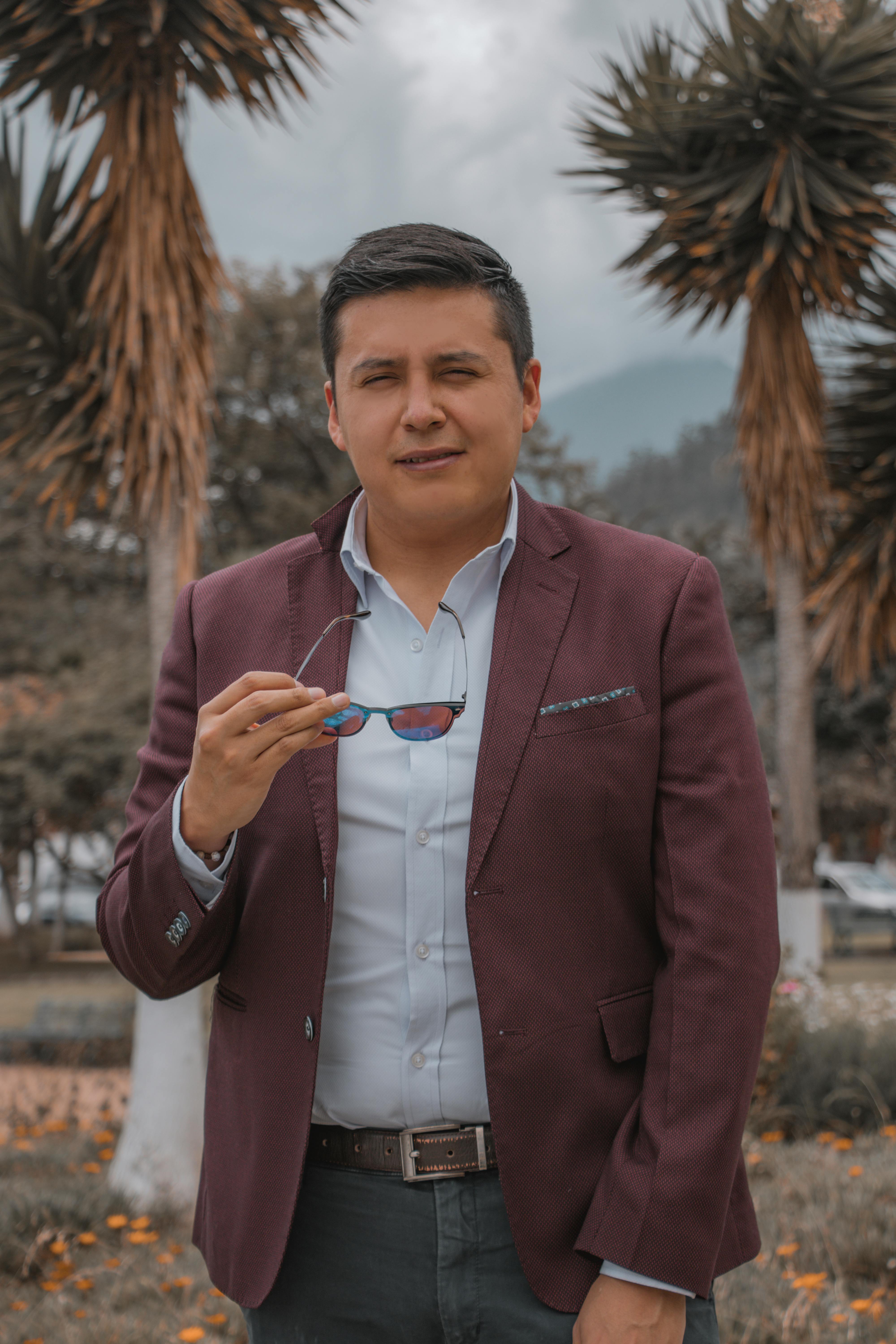 Man in a burgundy blazer holding sunglasses poses outdoors. Fashionable and casual style.