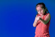 Indian little girl in casual clothes standing near blue wall and looking at bracelet on hand
