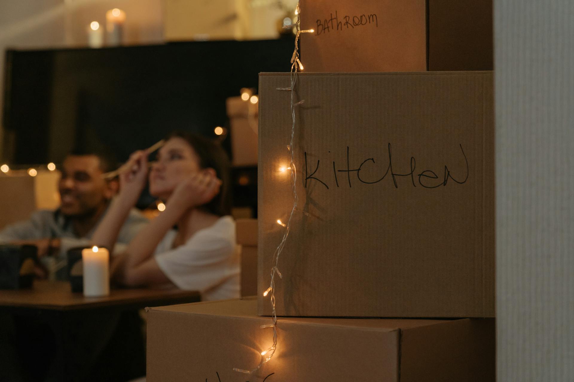 A couple relaxing among moving boxes in a softly lit room with candles and string lights, creating a cozy atmosphere.