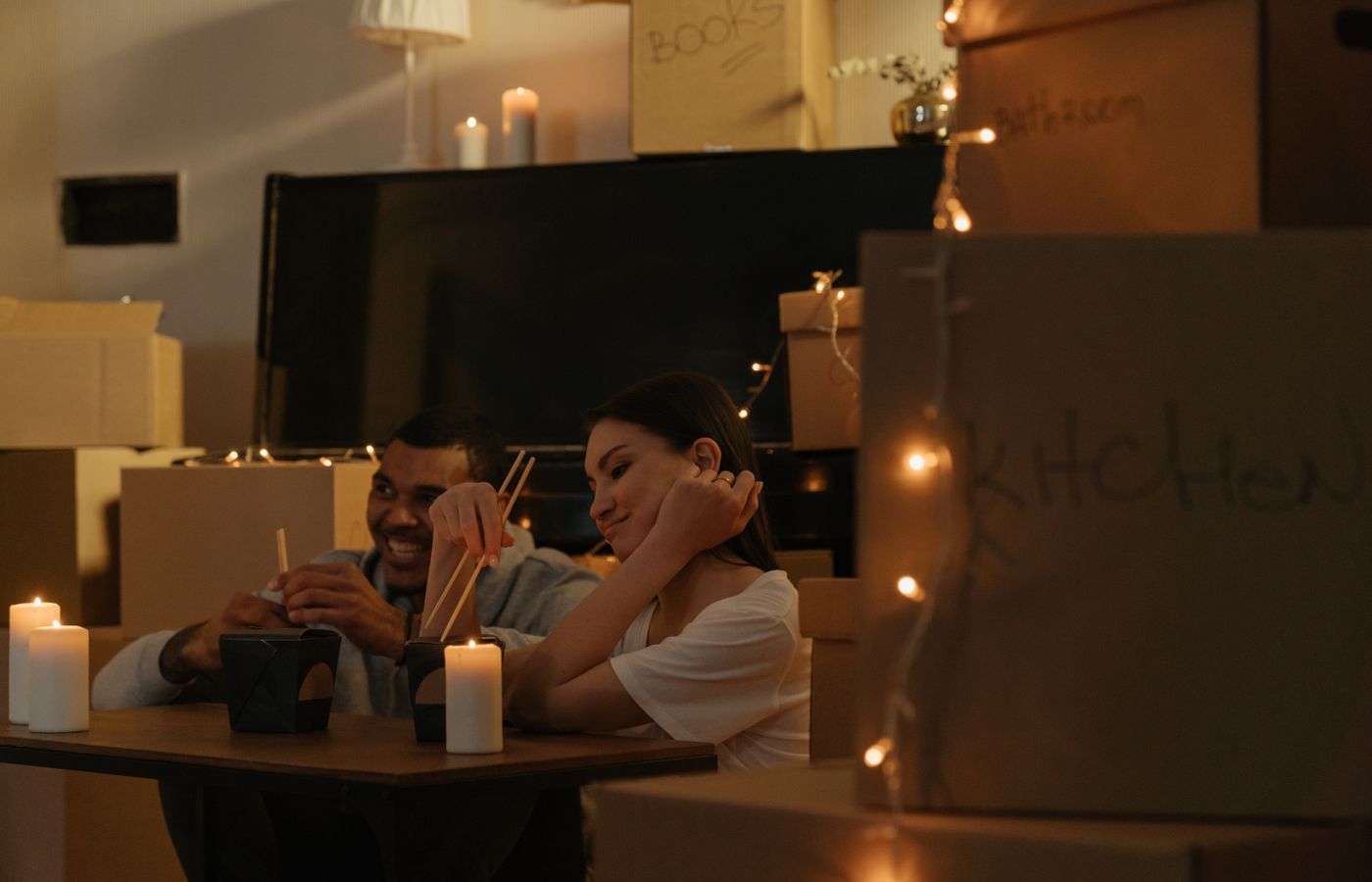 A couple enjoys a candlelit dinner with chopsticks surrounded by moving boxes in a cozy setting.