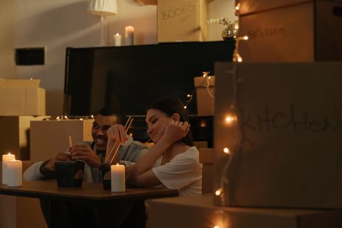 Free A couple enjoys a candlelit dinner with chopsticks surrounded by moving boxes in a cozy setting. Stock Photo