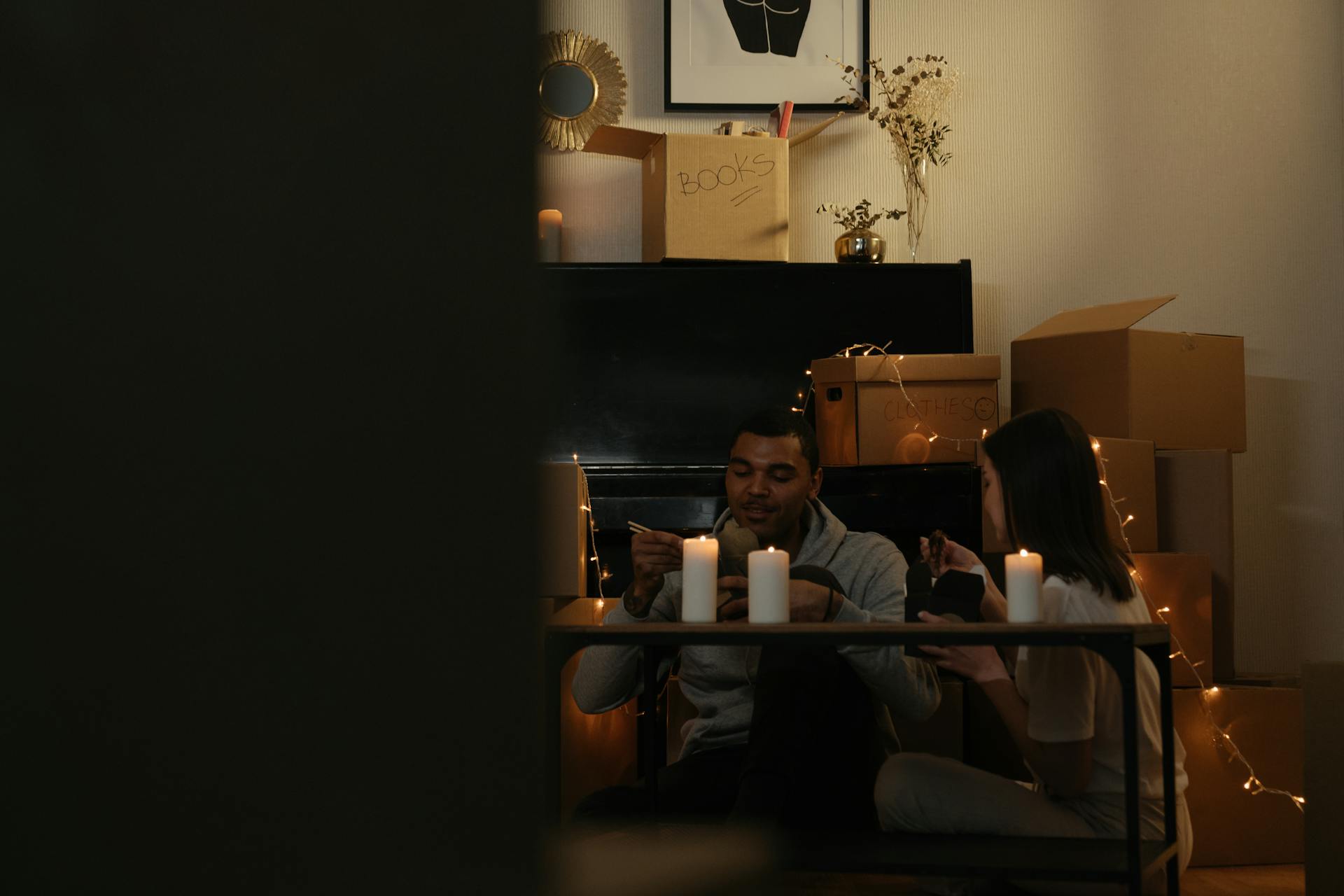 Couple enjoying a meal by candlelight surrounded by moving boxes in a cozy home setting.