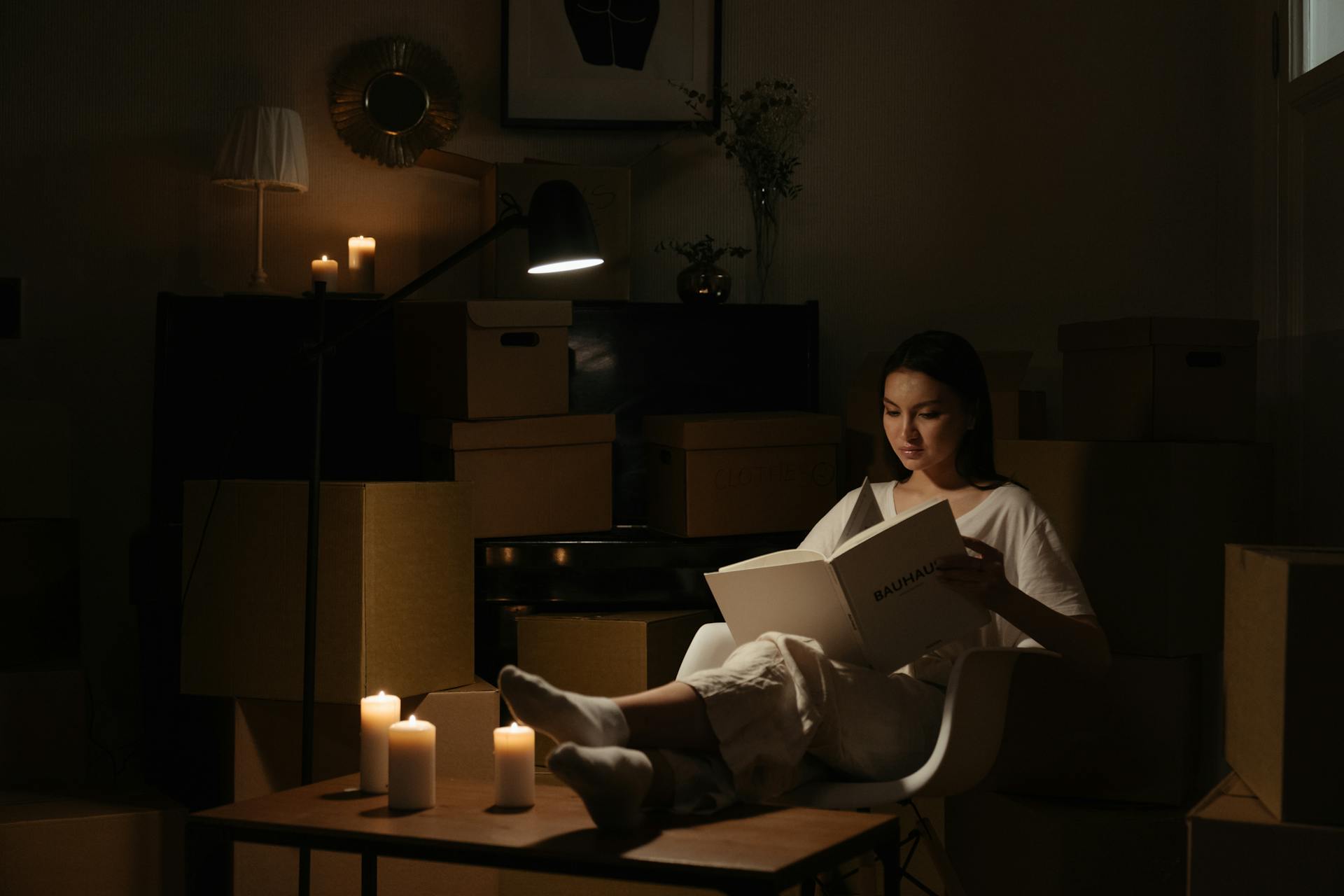 A woman relaxes reading a book by candlelight among moving boxes, creating a serene ambiance.